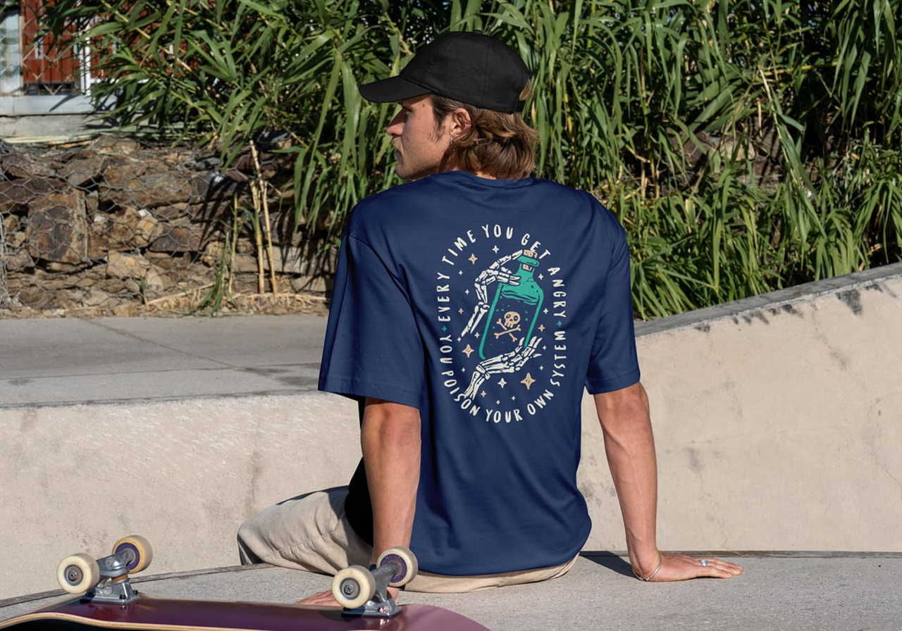 A man wearing a navy shirt with a skeleton hand and bottle graphic sits on a ledge at a skate park.