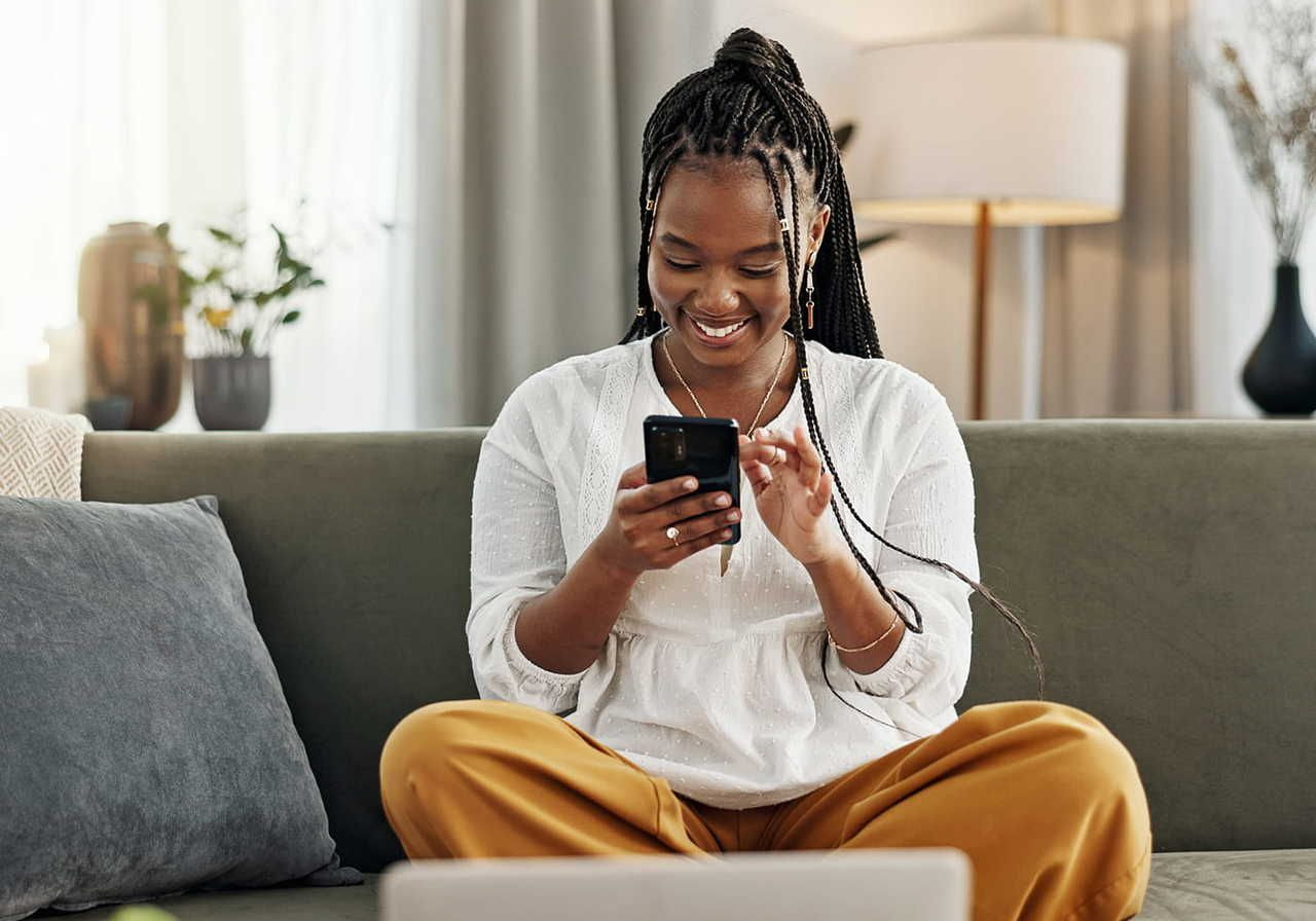 A woman with braided hair sits on a couch, smiling while using a smartphone. She wears a white top and yellow pants.