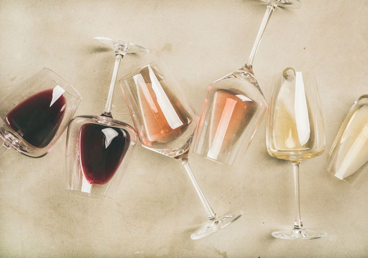 Wine glasses filled with red, rosé, and white wine lie on their sides on a beige stone surface, seen from above.