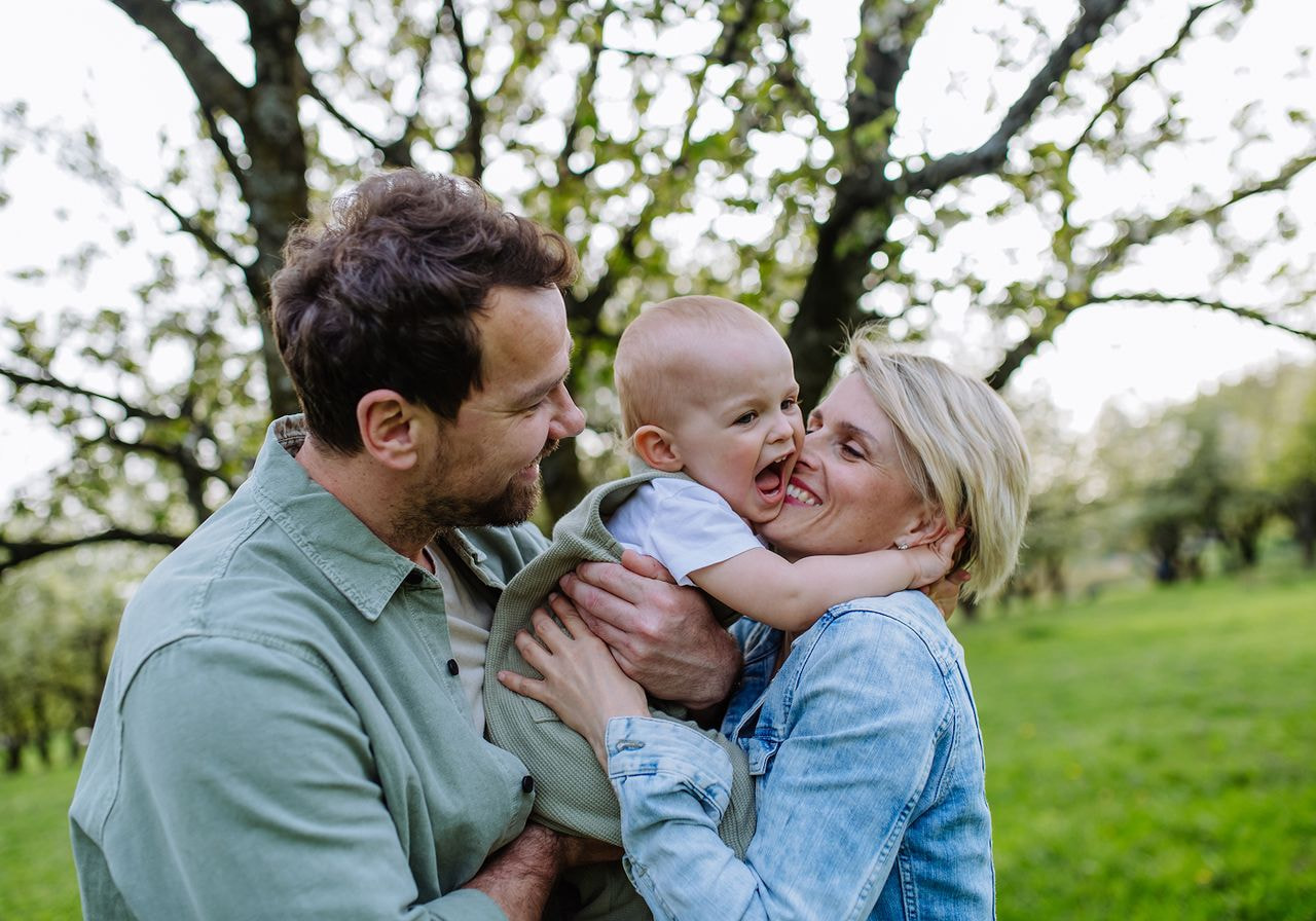 A man and woman hold their baby in an orchard, surrounded by trees laden with fruit, enjoying a sunny day together.