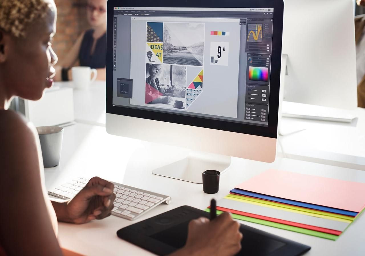 A woman works on graphic design at a computer, using a tablet. The screen displays colorful geometric layouts.