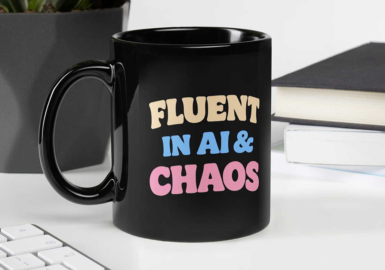 Black mug with bold text: "Fluent in AI & Chaos" in cream, blue, and pink. Positioned near a keyboard, books, and a plant on a desk.