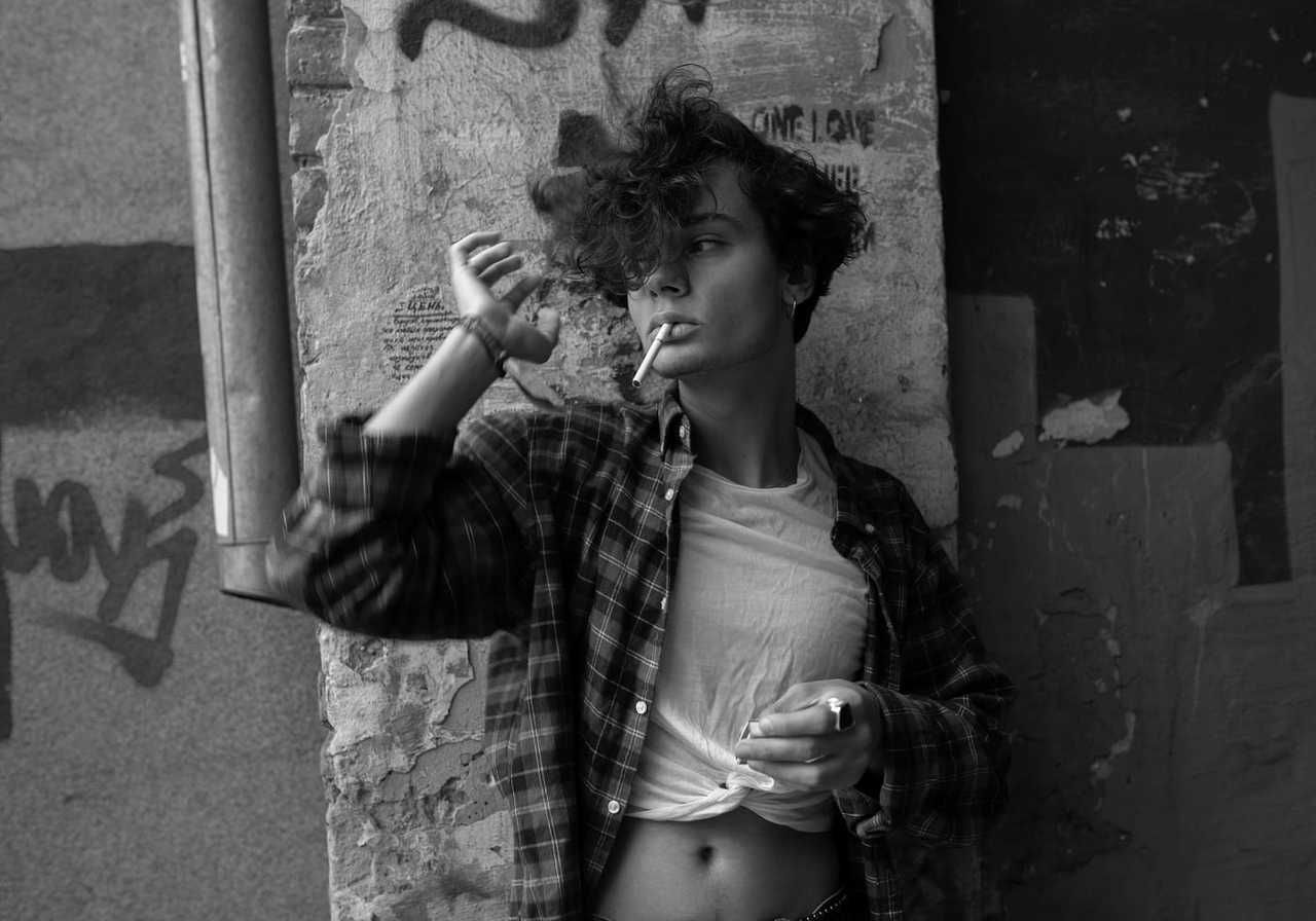 Black and white photo of a woman in a flannel shirt with tousled hair leaning against a graffiti wall, holding a cigarette.