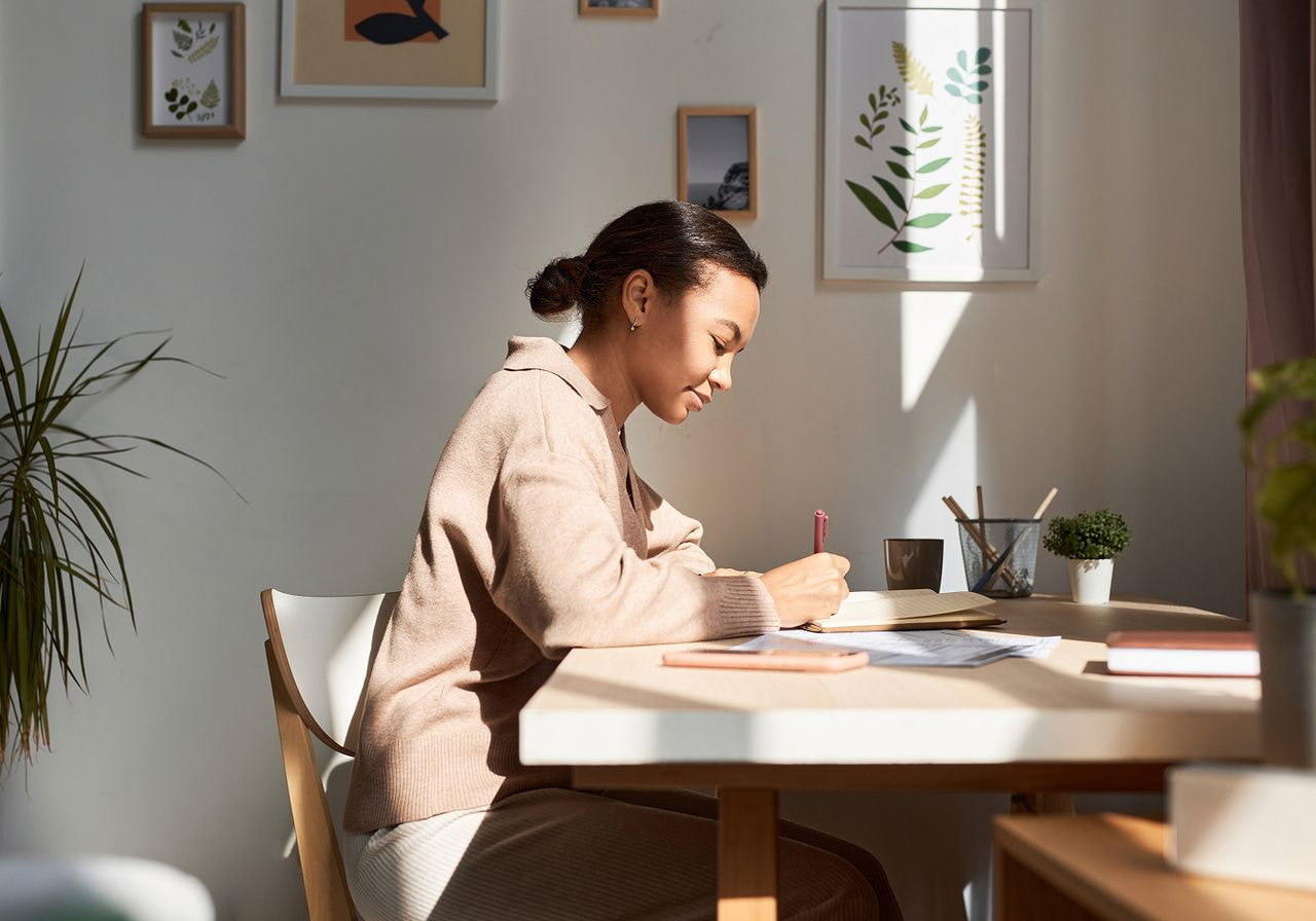 A woman writes in a journal at a sunlit wooden desk in a calm home office decorated with botanical prints.