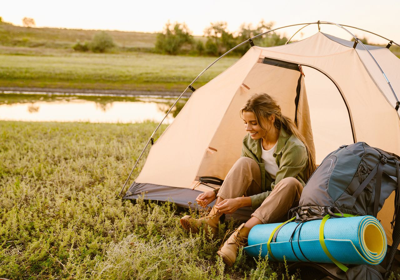 A woman sits in front of a tent, resting beside her backpack in a natural outdoor setting.