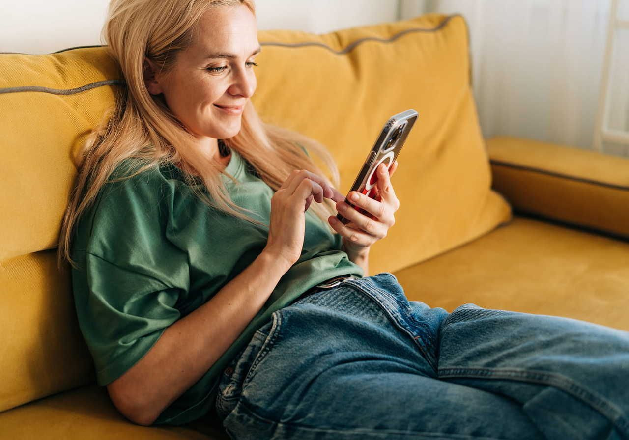 A smiling blonde woman in a green shirt and blue jeans, sitting on a yellow couch, with a smartphone in her hands.