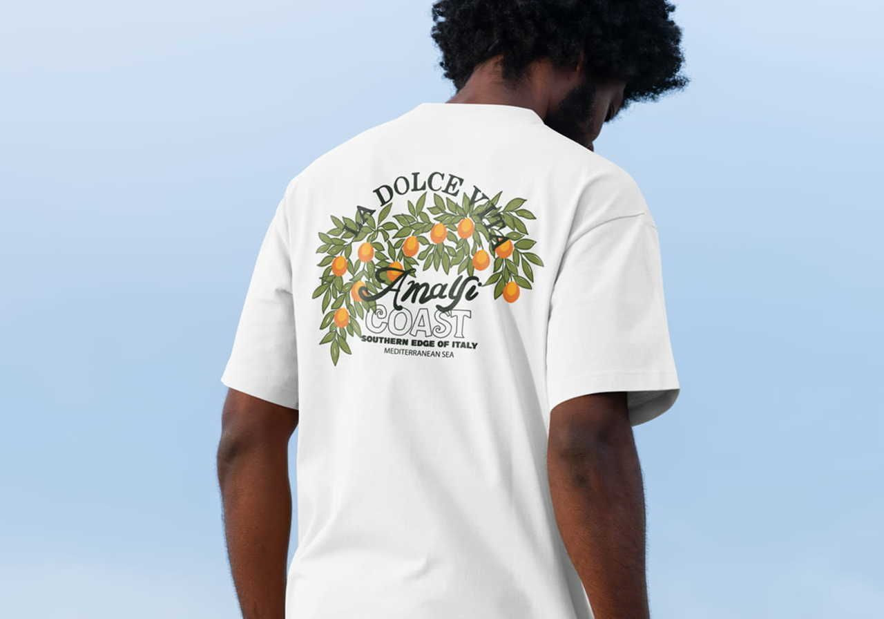Man wearing a white t-shirt with "La Dolce Vita Amalfi Coast" and orange branches design on the back, against a clear blue sky.