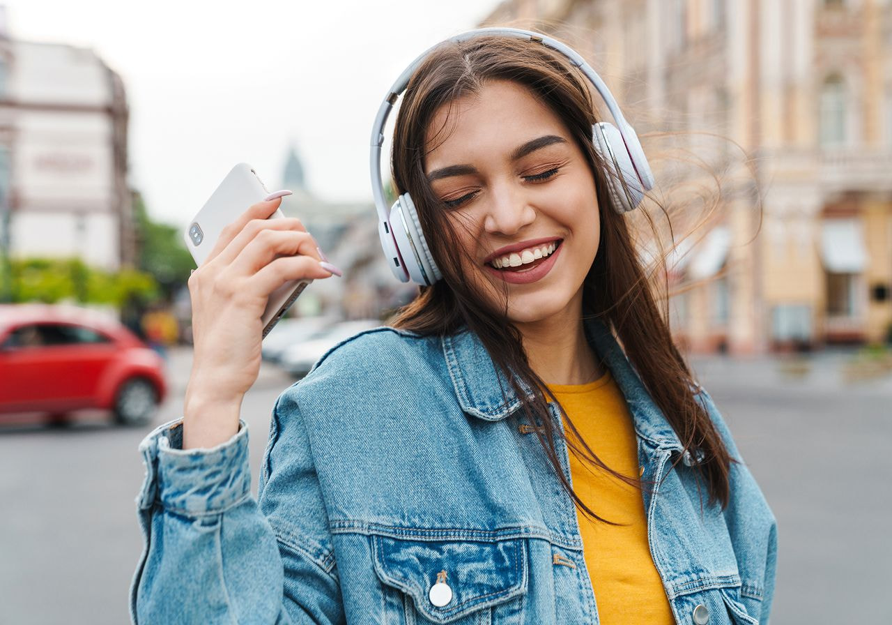 Smiling woman wearing headphones holding her phone while walking outdoors.