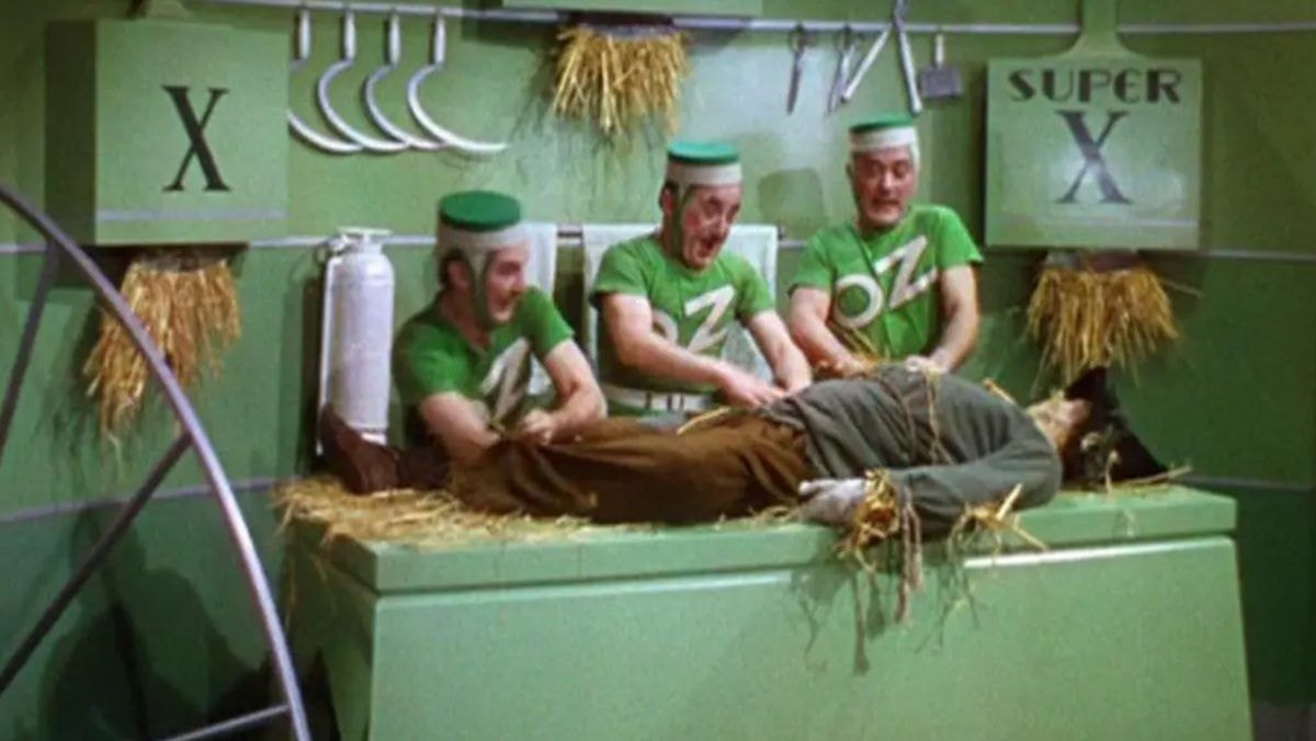 Three men in green uniforms excitedly groom a man lying on a table with straw decor