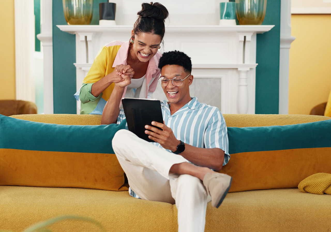 A joyful couple on a yellow and teal sofa, smiling at a tablet. Set in a bright, stylish living room.