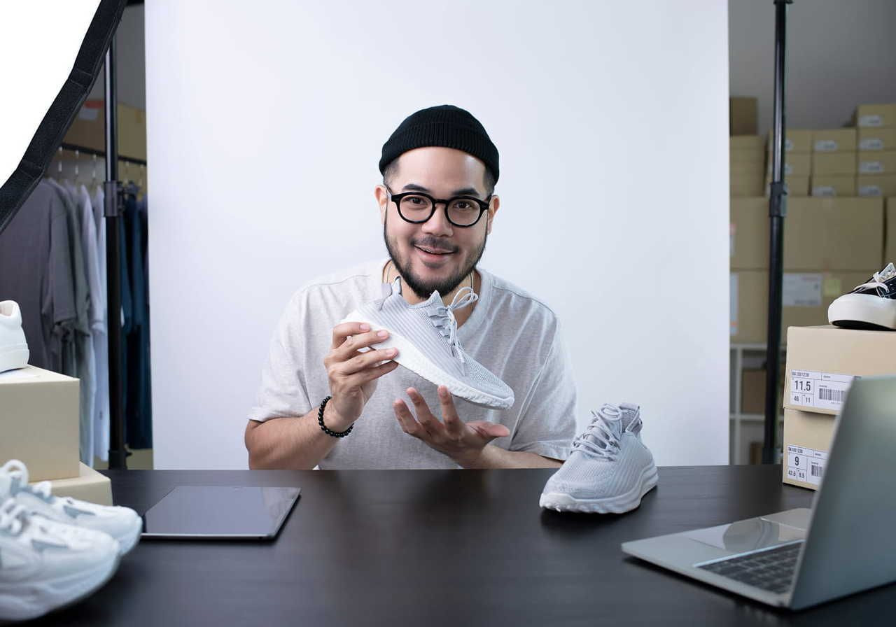 A man wearing glasses and a beanie holds a sneaker while seated at a table with shoes and a laptop.
