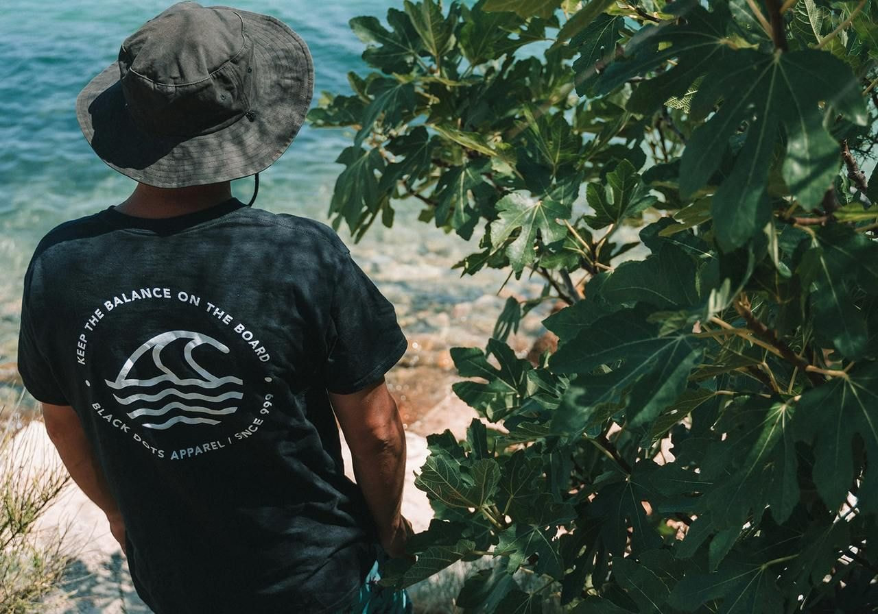 A man is standing in front of a sea coast line, wearing a blue t-shirt with a large back print.