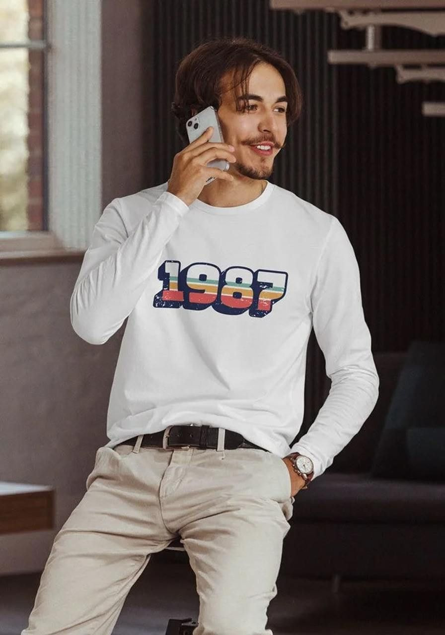 A man in a white long-sleeve shirt with "1987" in bold, colorful font stands near a window, smiling while talking on a phone.