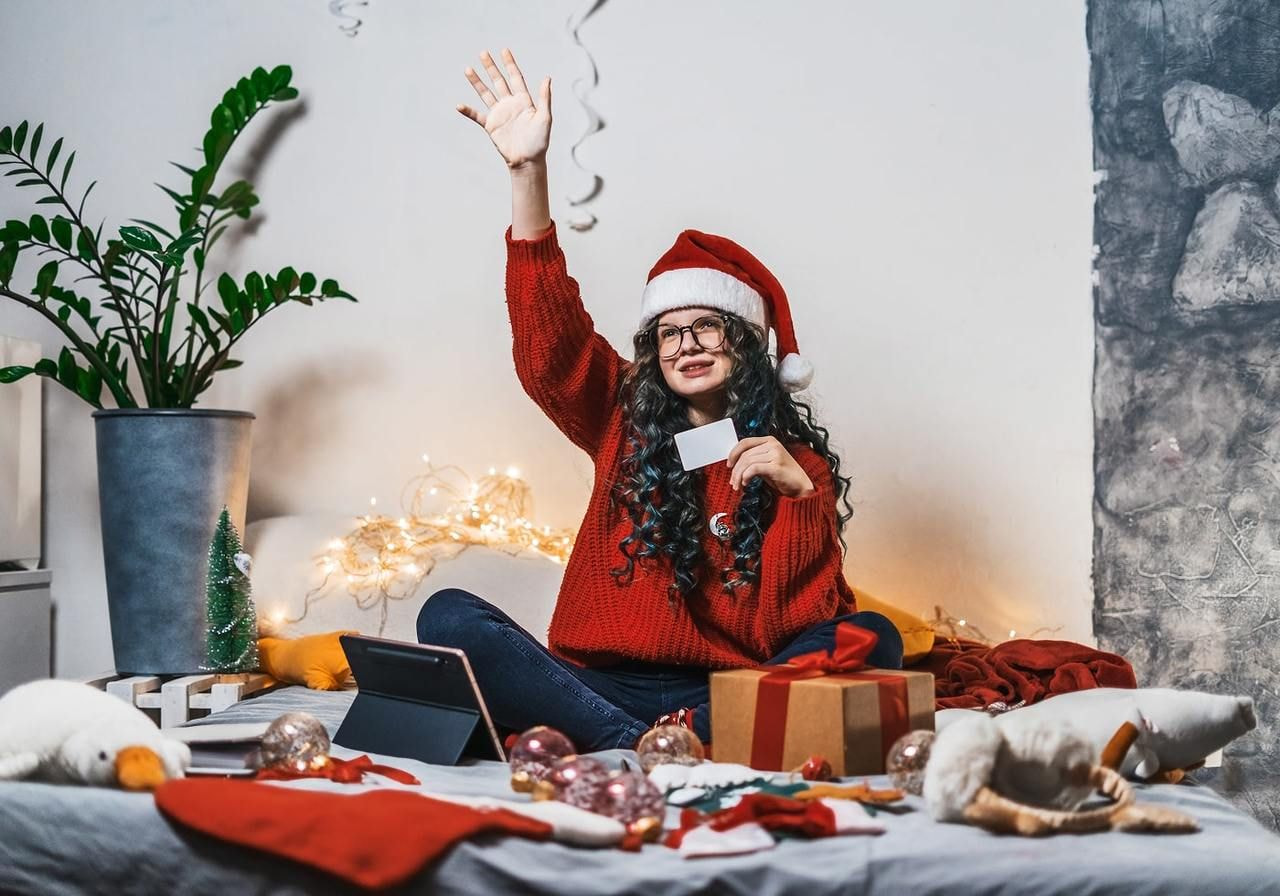 A young woman in a Santa hat and red sweater sitting on a bed with festive decorations, smiling, raising her hand.