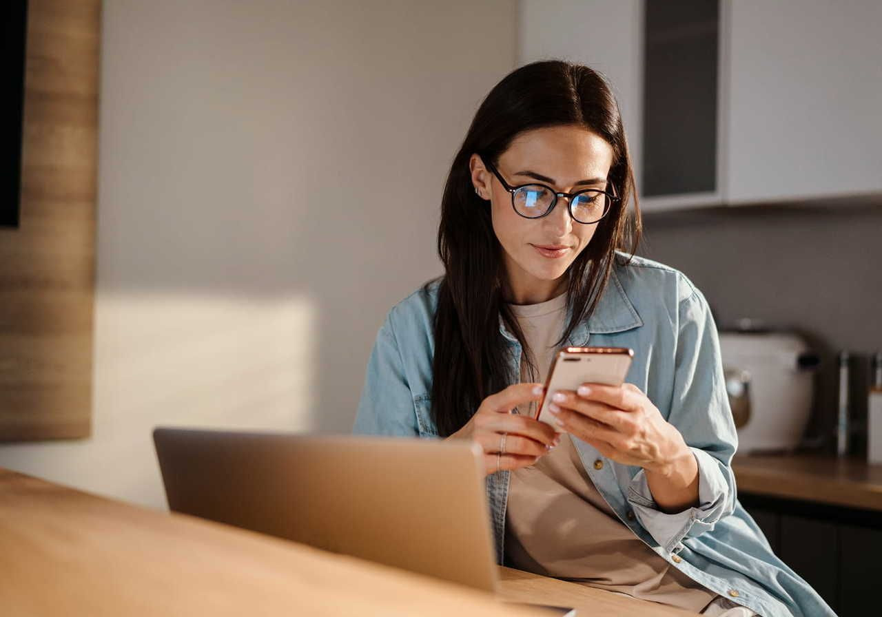 A woman wearing glasses is sitting at a wooden table, looking at her smartphone with a laptop in front of her.
