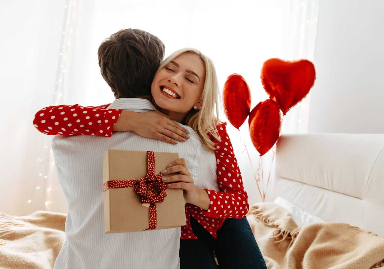 A woman in a red polka dot shirt joyfully hugs a person while holding a gift with red ribbon. Red heart balloons float nearby.