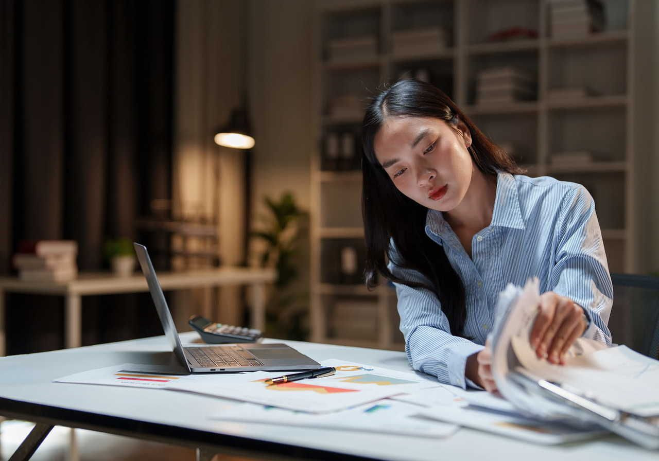 A woman in a blue shirt works at a tidy desk in a dimly lit office, focused on reviewing documents. A laptop, papers, and a pen are visible.