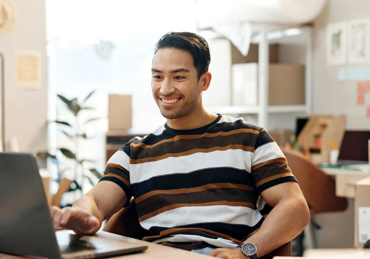 A smiling man in a striped shirt is sitting at a laptop, searching: is selling on Etsy worth it?