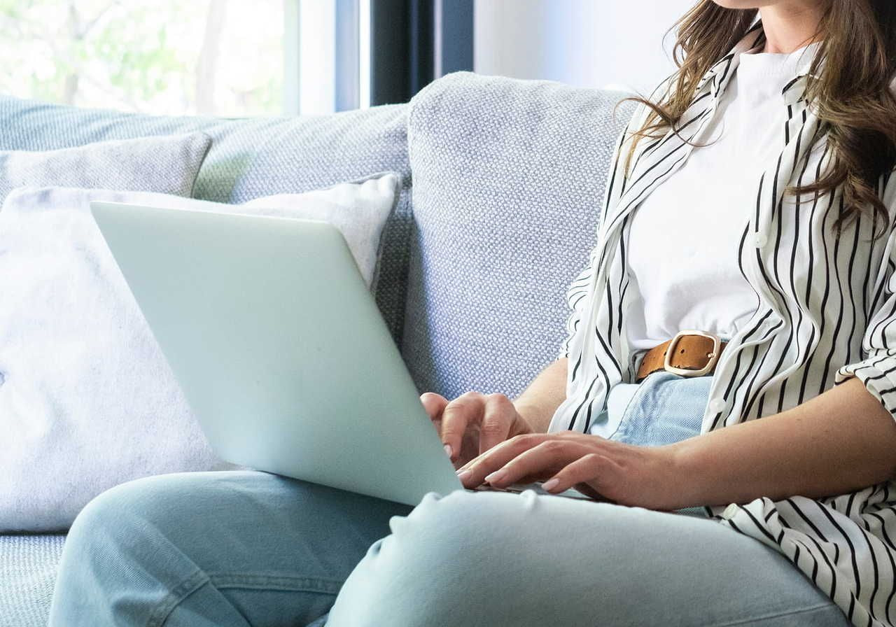 A person sitting on a couch, wearing a striped shirt and light jeans, using a laptop.