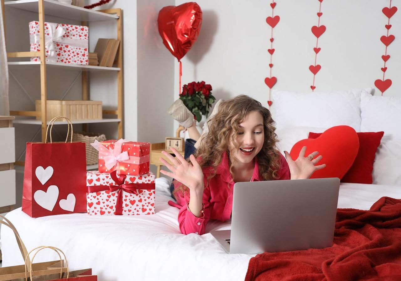 A young woman on a bed, excitedly looking at a laptop, searching for Valentine’s Day marketing ideas, with gifts in the background.