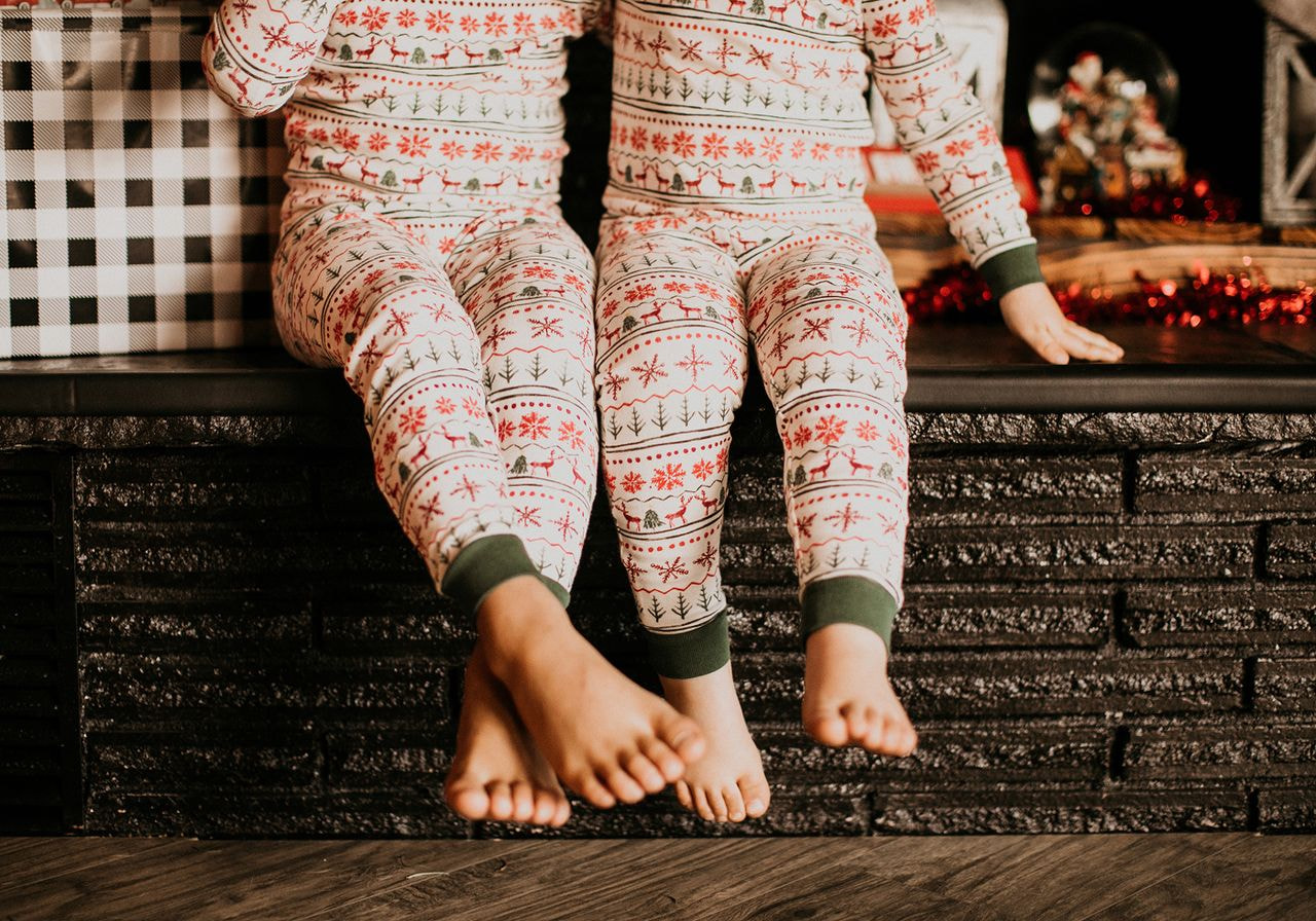 Two children in matching Fair Isle Christmas pajamas with reindeer and snowflake prints sit barefoot on a dark brick ledge beside wrapped gifts.