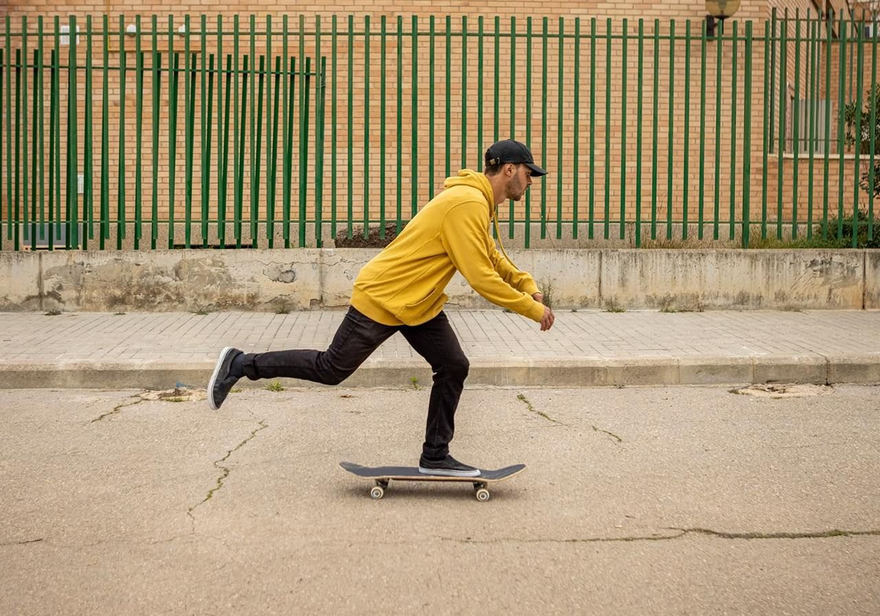 A man in a yellow hoodie and black cap skateboards on a street beside a tall green fence.
