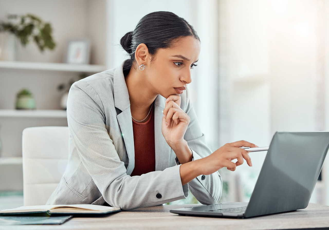 A woman in a blazer sits at a desk, looking thoughtfully at her laptop while holding a pen.