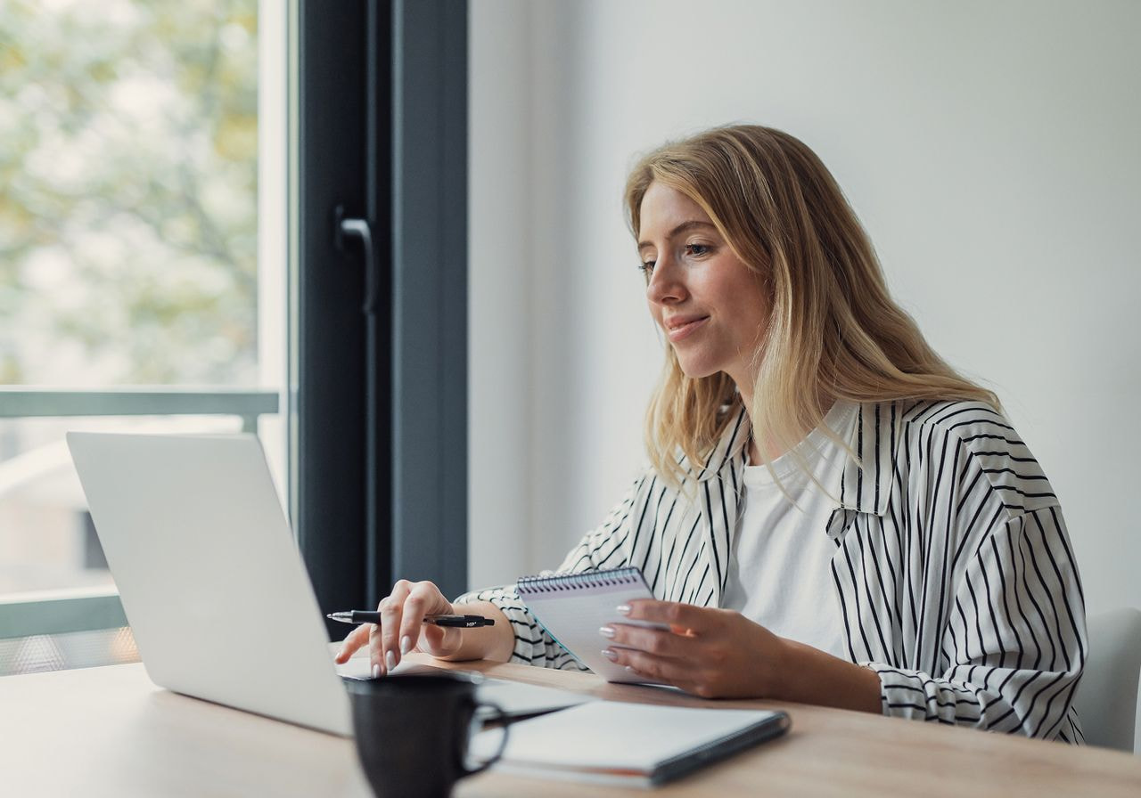 A woman in a striped shirt reviews notes and works on a laptop at a desk by a window.