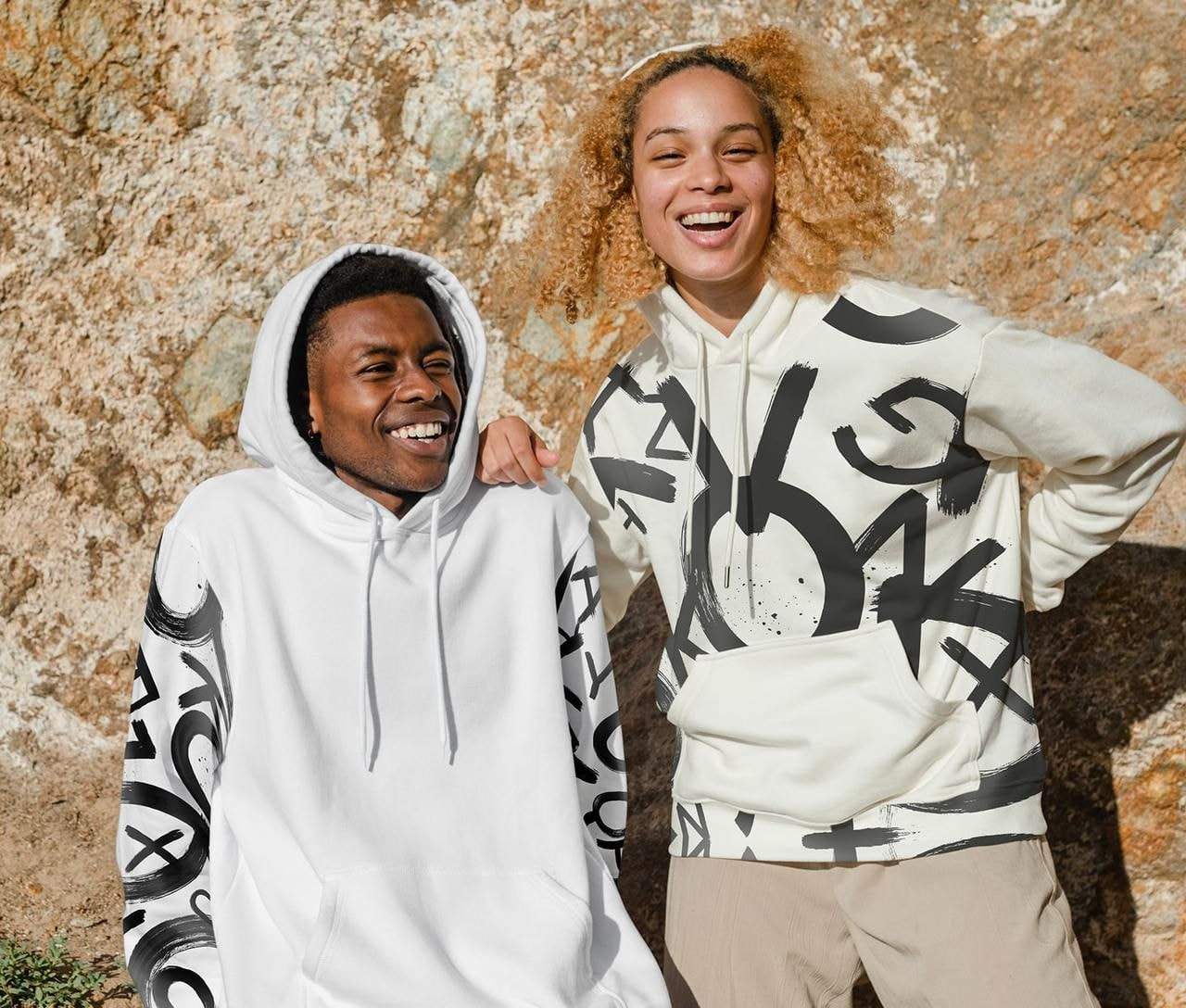 Two people smile warmly, wearing graphic hoodies with abstract designs, against a textured stone wall.