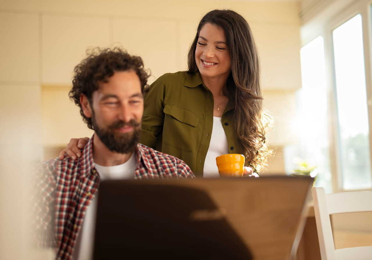 A smiling woman in casual attire holds a yellow mug, standing behind a bearded man working on a laptop.