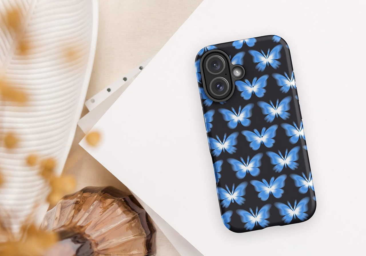 A smartphone with a black case featuring blue butterfly patterns lies on a white surface beside a spiral notebook and decorative items.