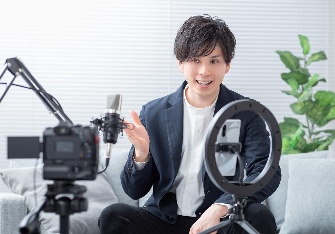 A man in a suit sits on a couch, speaking into a microphone with a camera and ring light setup.