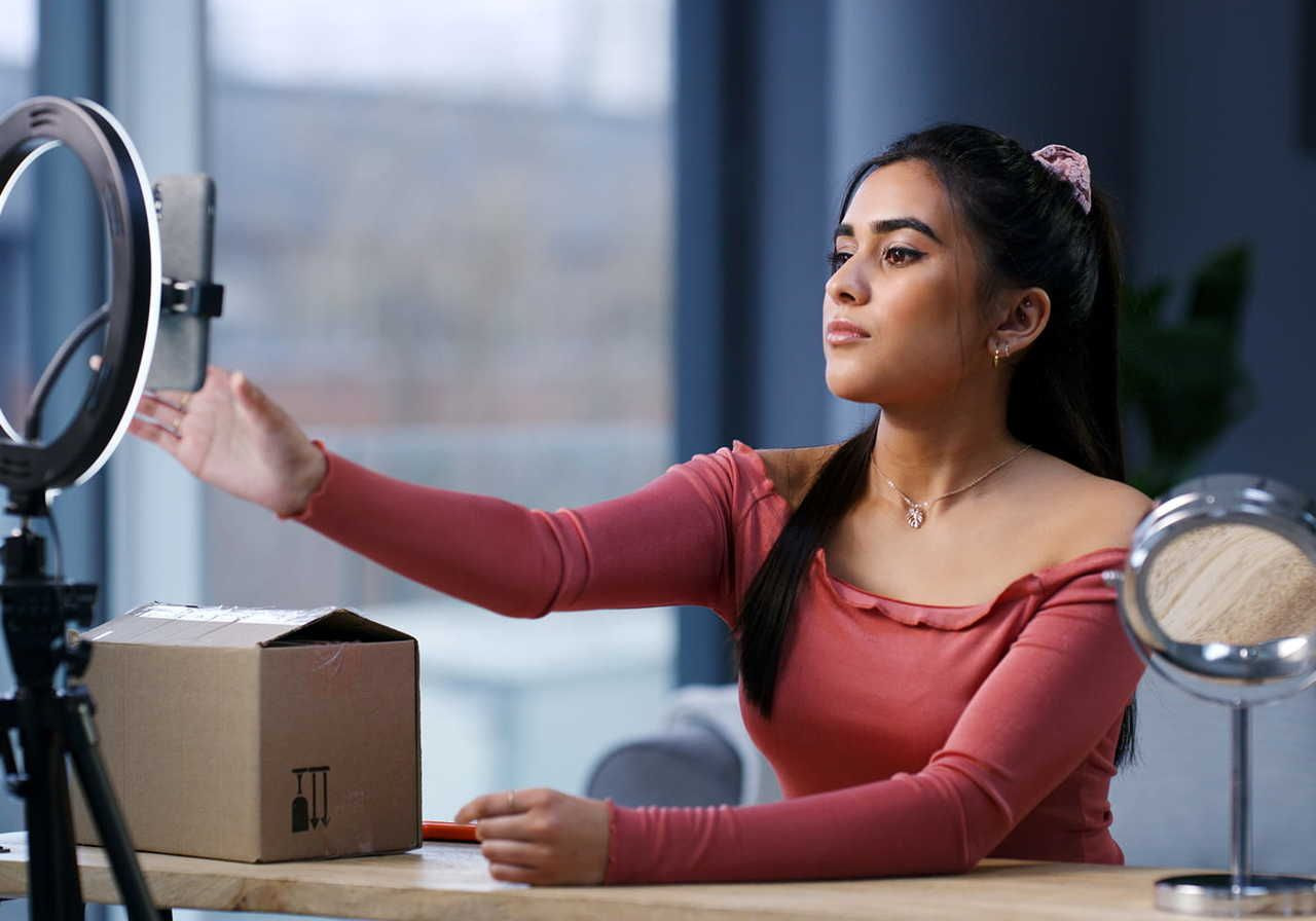 Young woman filming herself at a table, adjusting a ring light with a smartphone. A makeup mirror and a cardboard box are on the table.