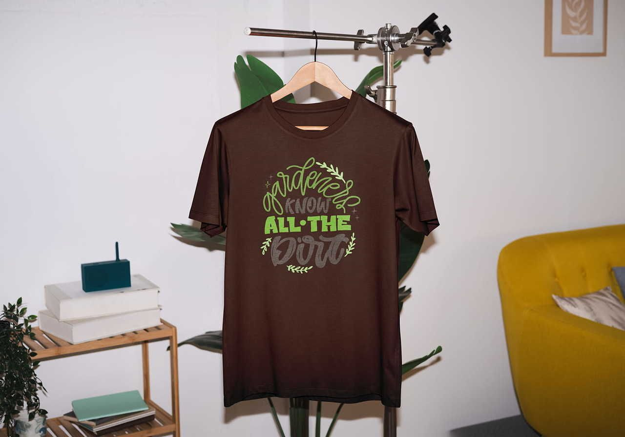 A brown t-shirt with green text "Gardeners Know All the Dirt" hangs on a rack in a room with a yellow sofa and plants.