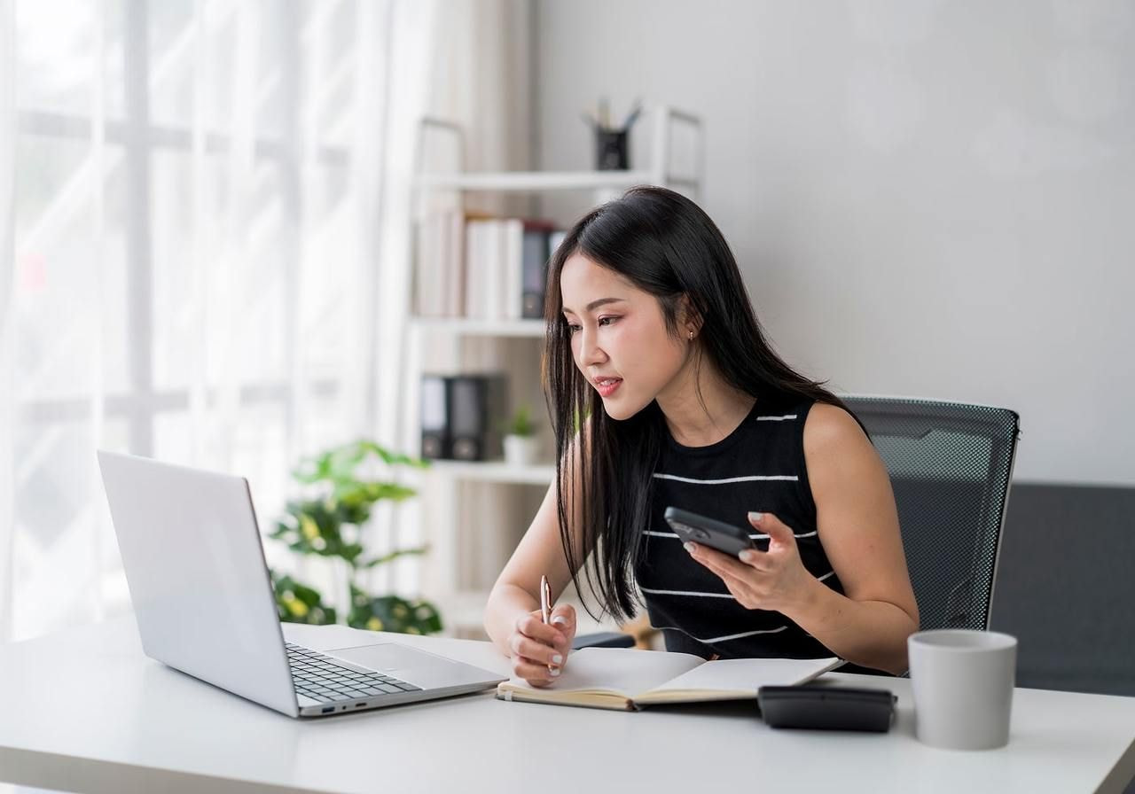 A young woman is making notes in a notebook while sitting in front of a laptop, holding a mobile phone.
