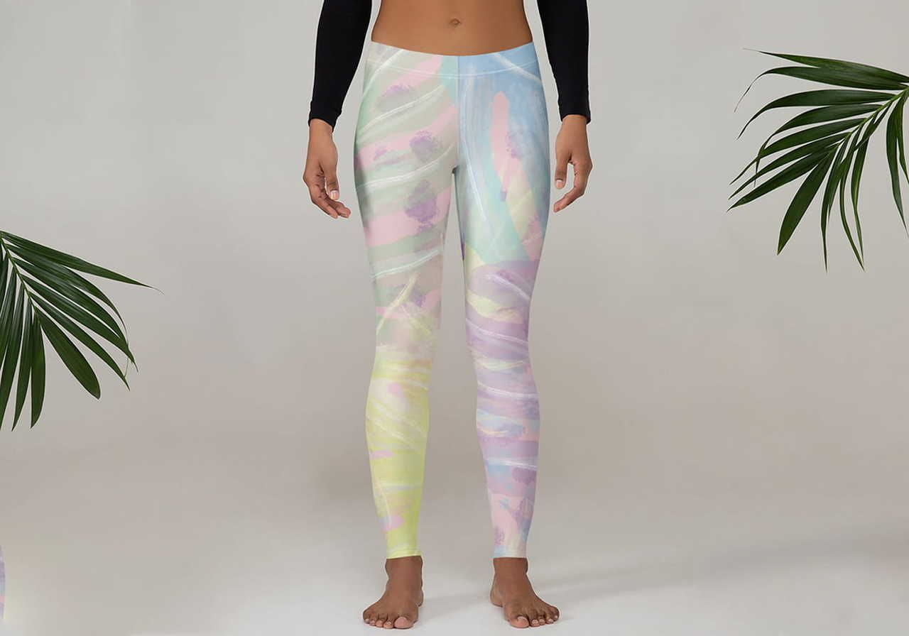 A woman wearing colorful, pastel-patterned leggings and a black long-sleeve top stands barefoot on a light background, flanked by green leaves.