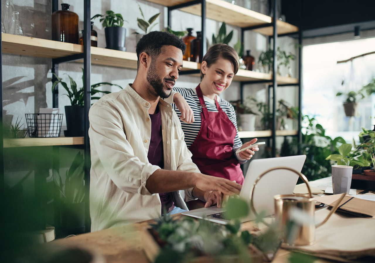A man working on a laptop at a table with a woman standing beside him in a plant-filled workspace.