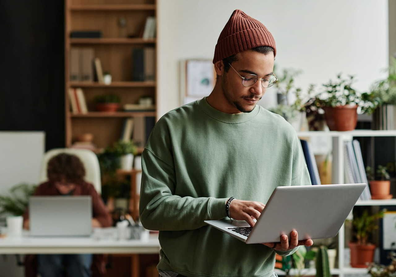 A man in a green sweater and a beanie is working on a laptop, standing in an office with plants and a person in the background using a computer.