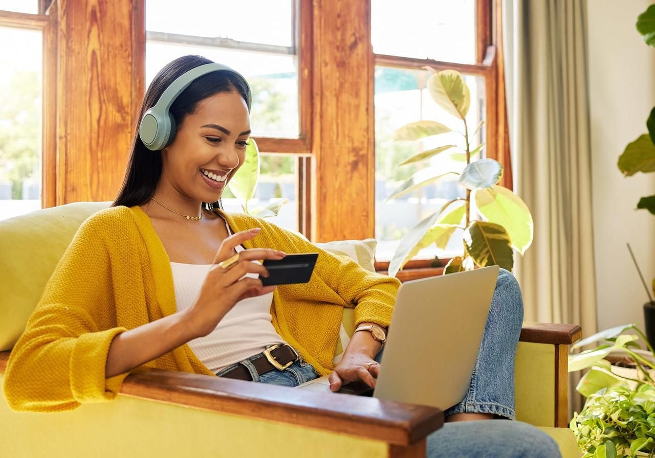 A woman in a yellow sweater, wearing headphones, smiles while viewing a laptop and holding a credit card.