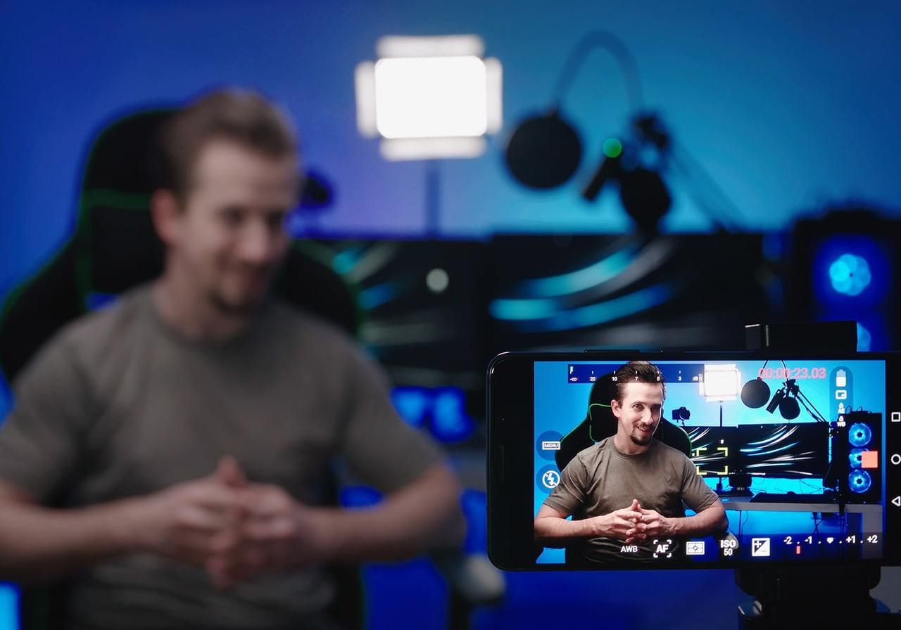 A man in blurry foreground sits with hands clasped, facing a smartphone displaying him in focus.