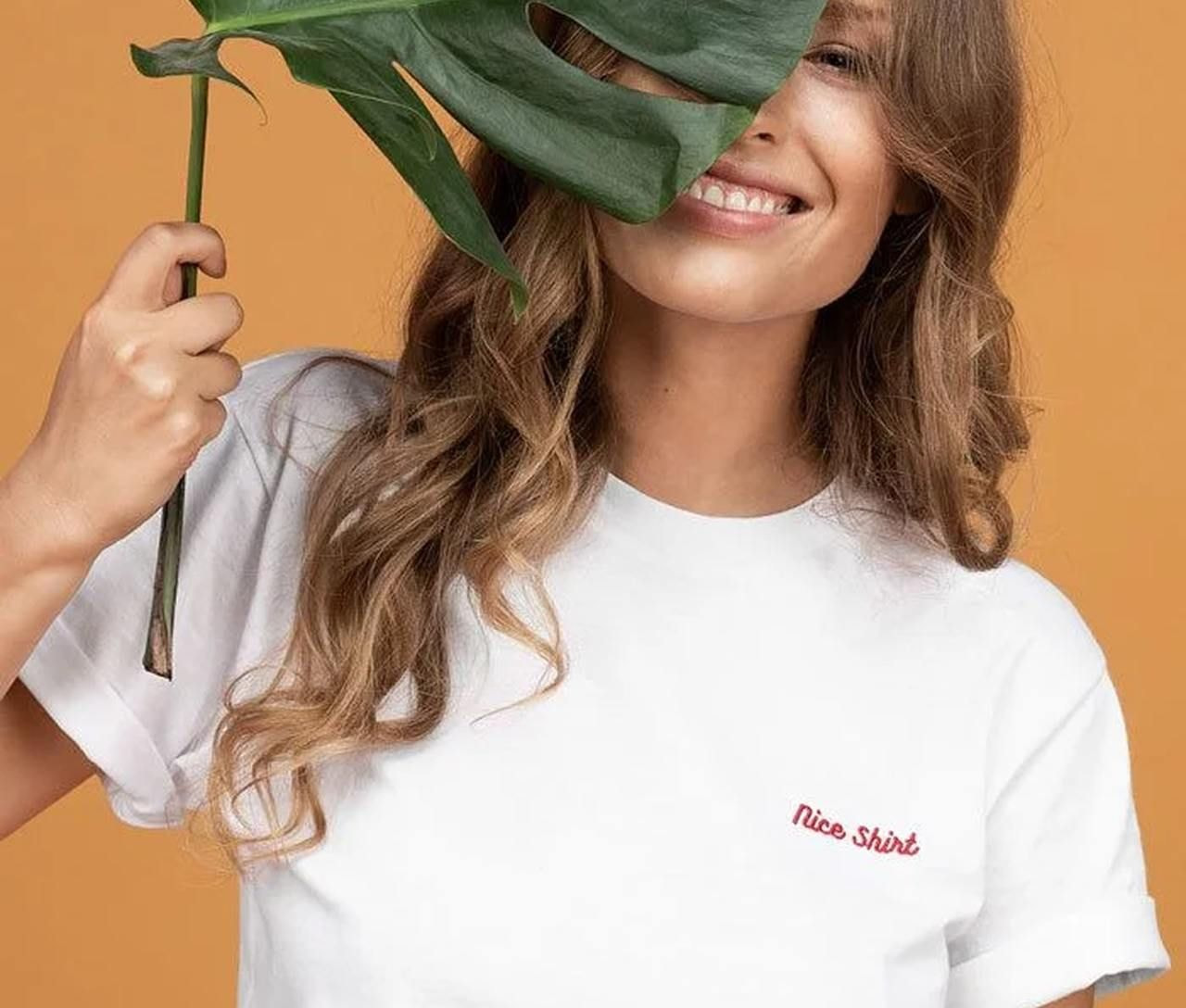 A smiling woman holds a large green leaf partially covering her face. She wears a white t-shirt with "Nice Shirt" text embroidered.
