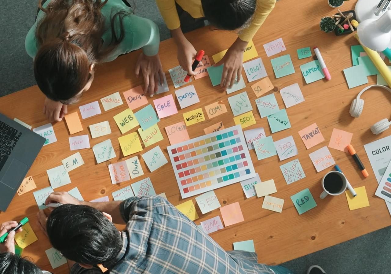 A group of people collaborates at a wooden table filled with colorful sticky notes, a color chart, and markers.