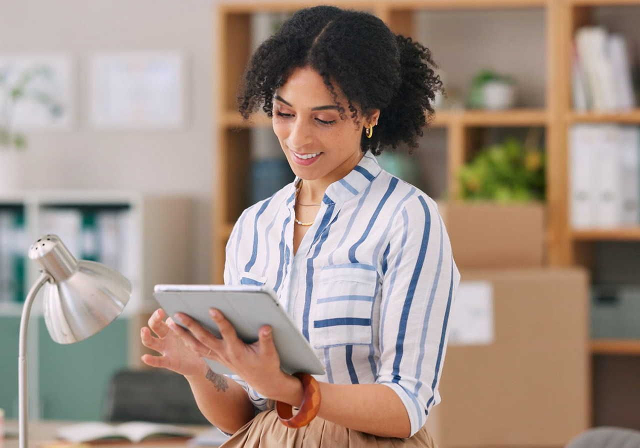 A woman with curly hair is standing in an office setting, holding a tablet and smiling while looking at it. She is wearing a striped shirt.
