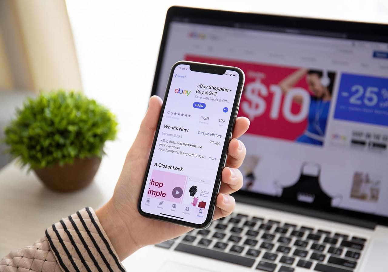 A person holds a phone displaying the eBay app screen in front of a laptop showing the eBay homepage.