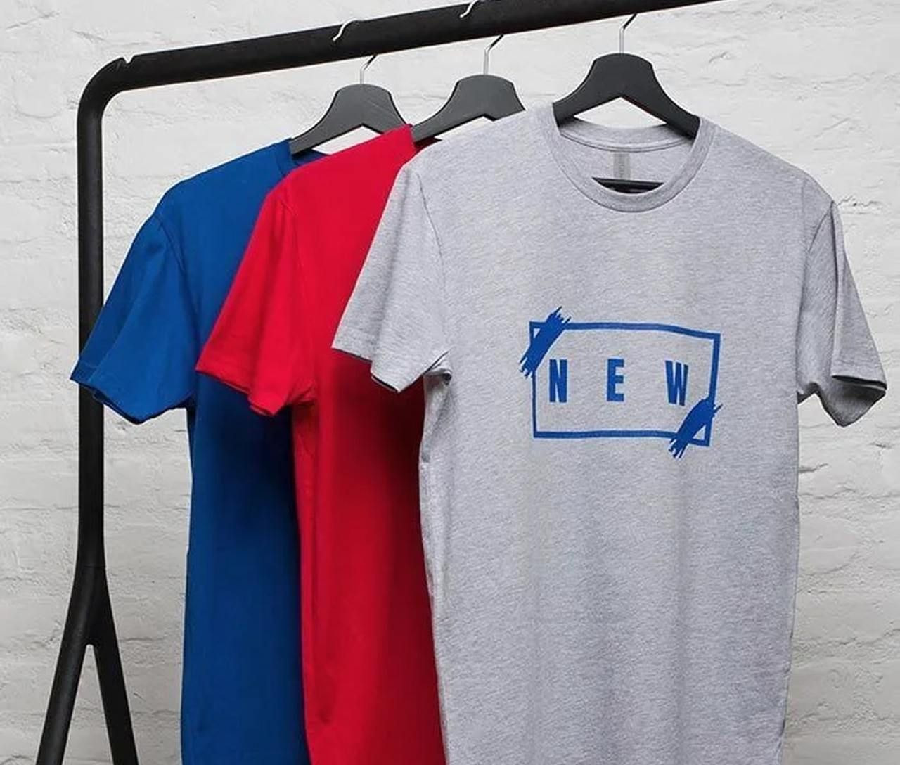 Three t-shirts on hangers: blue, red, and gray. The gray shirt features a blue design with the word "NEW."