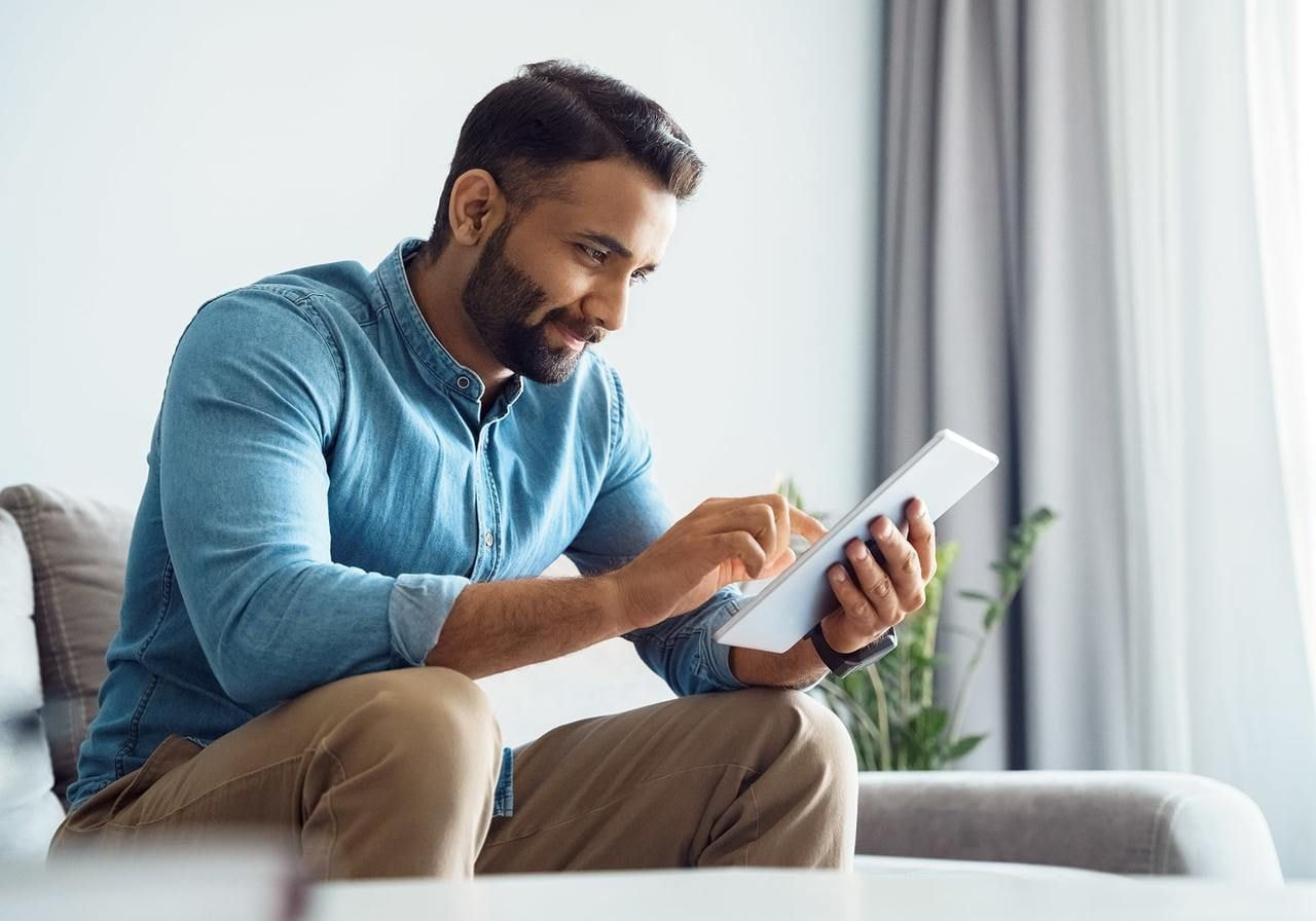 A man in a blue shirt sits on a sofa, smiling as he uses a tablet. Bright room with a potted plant and curtains.