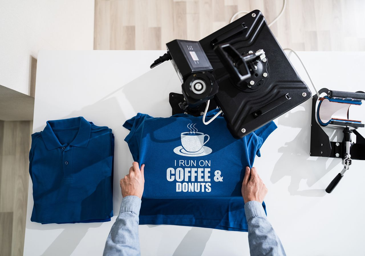 A person holds a blue shirt featuring a coffee and doughnut design, showcasing a casual and playful style.