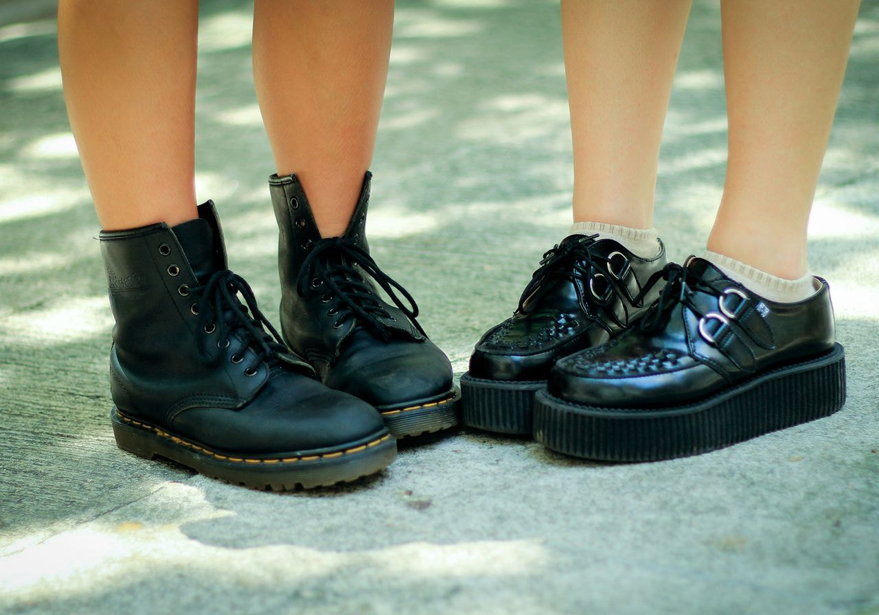 Close-up of two pairs of legs wearing black footwear: one in lace-up combat boots, the other in platform shoes, set on a sunlit concrete path.