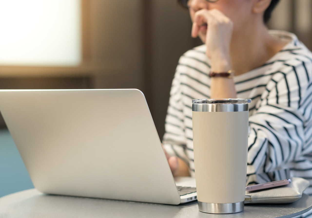 A person in a striped shirt works on a laptop at a table, searching for how to make custom tumblers. Travel mug and a smartphone are nearby.