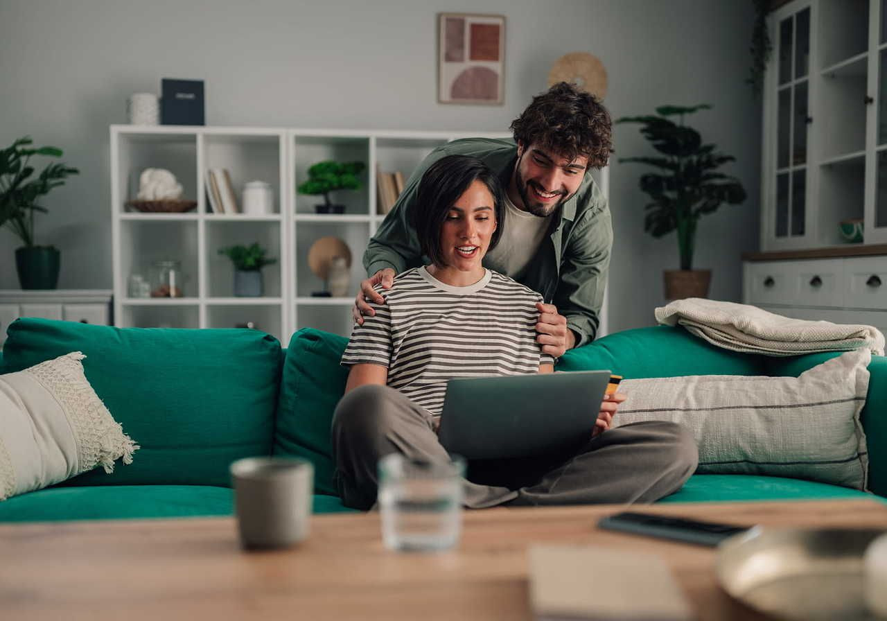 A smiling woman is sitting on a couch with a laptop, with a man patting her on the shoulders from the back.